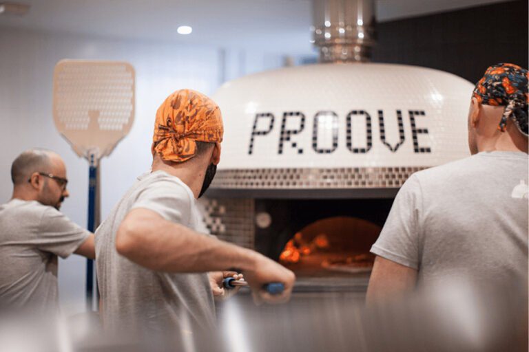 A large tiled pizza oven featuring the word 'Proove' within the tiles. A number of men are stood around it with pizza paddles, one is loading a pizza in to the oven.