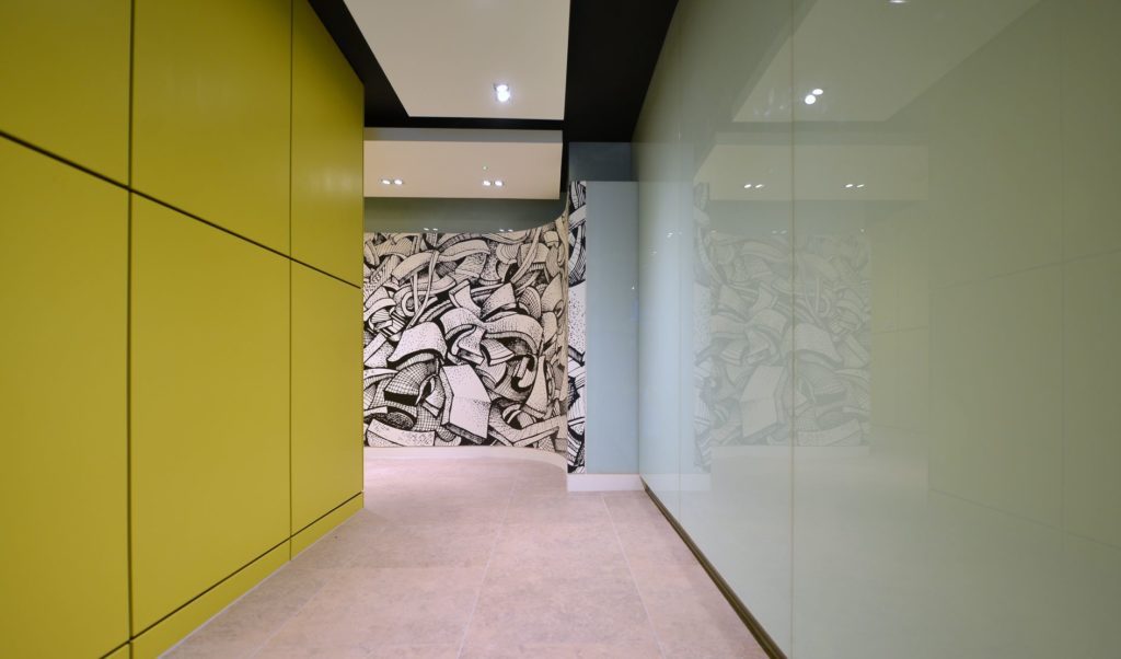 Corridor view with a large bright green panelled wall on one side, a reflective glass wall on the other side and a large black and white printed wall at the end of the corridor.