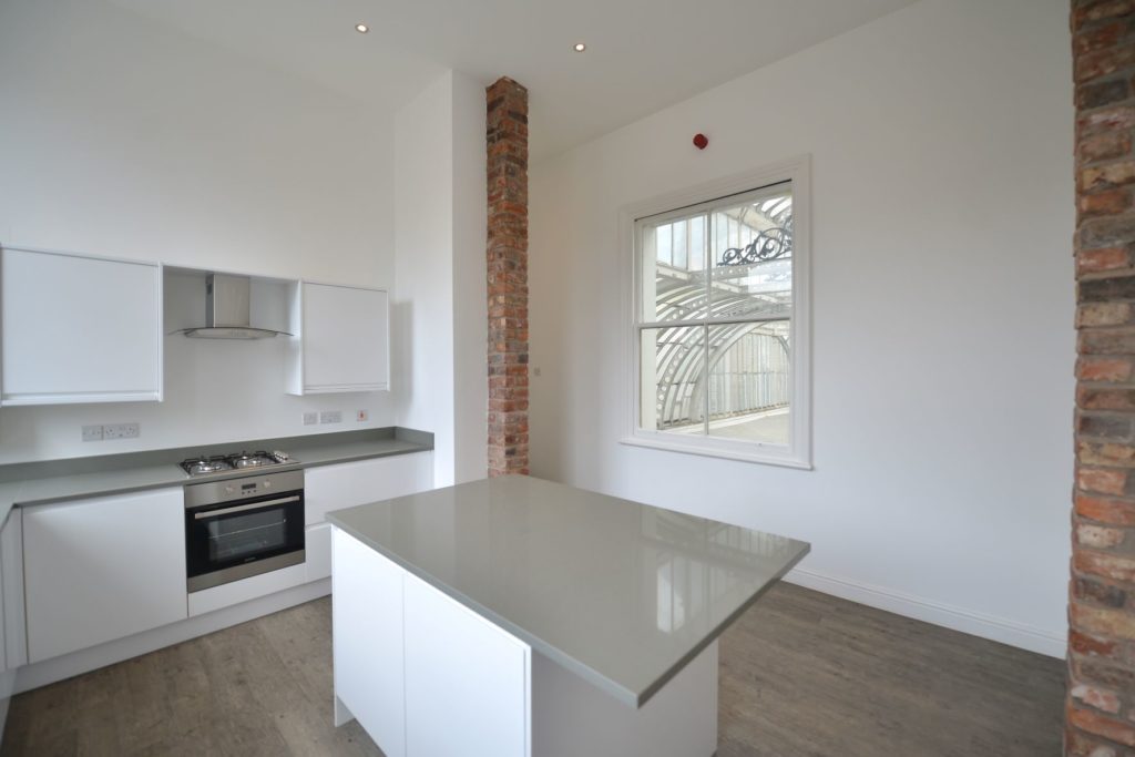 Internal image of a recently refurbished residential space showing a white and grey kitchen. There walls are white and there is some exposed brickwork.