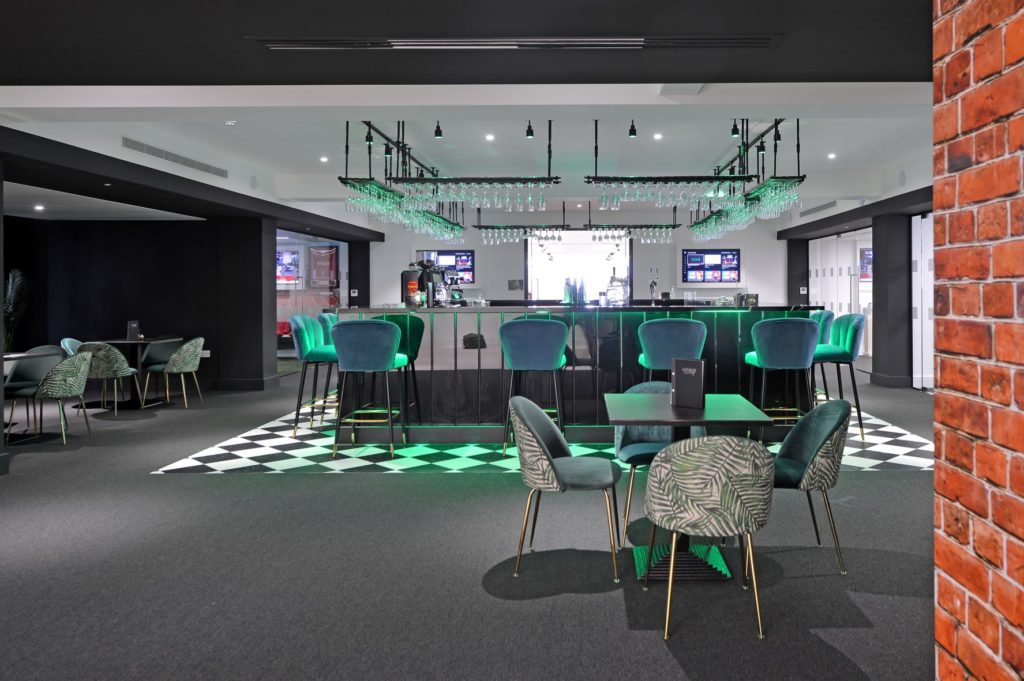 Bar area showing a long bar with tall stools around it, drinks glasses suspended from the ceiling above it and some smaller tables and chairs around it.