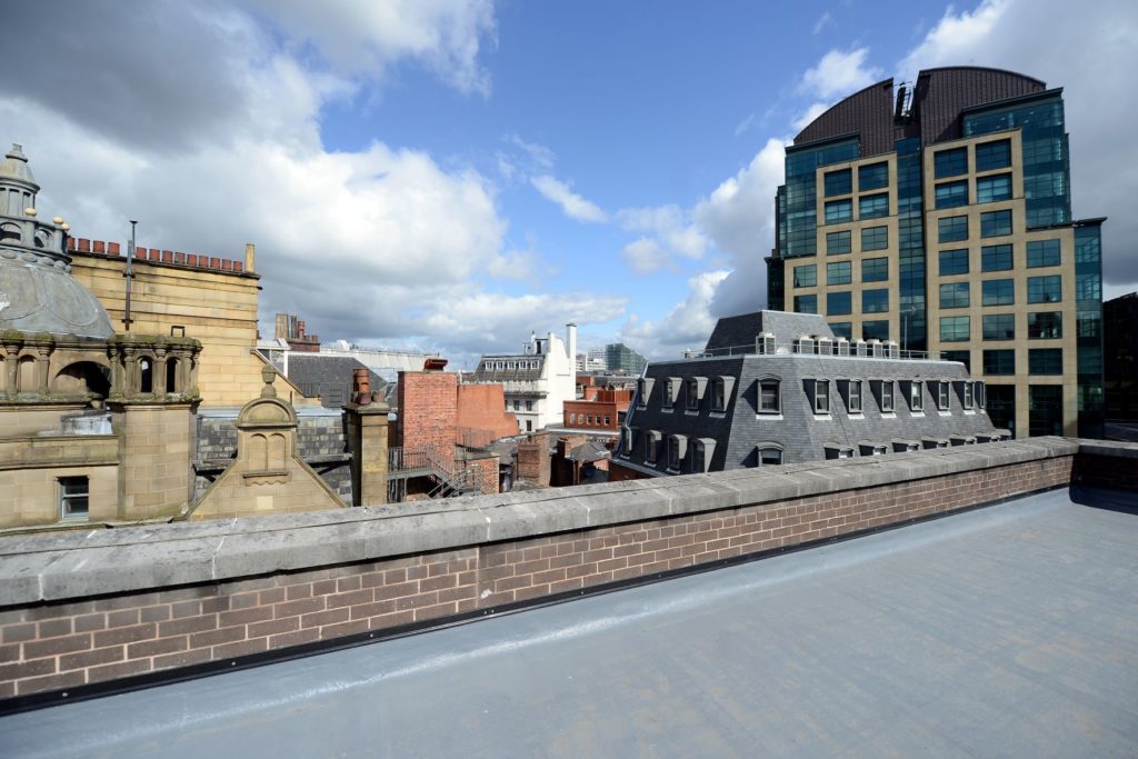 View over Manchester city centre from a rooftop. There are numerous buildings in view and the sky is blue but cloudy.