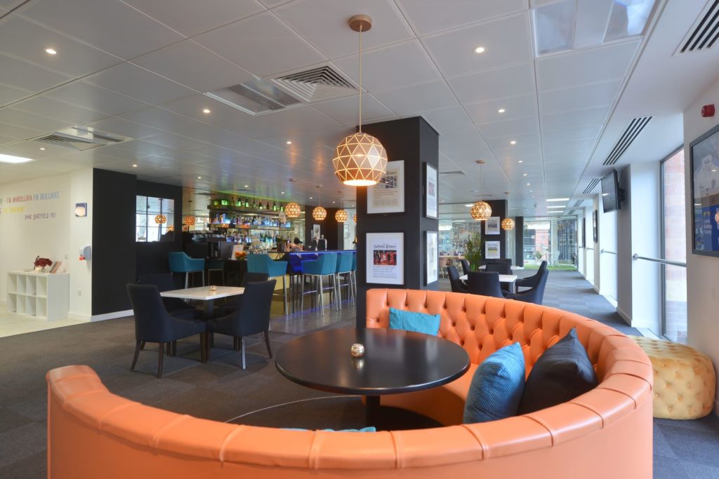 Lobby area featuring a large round orange booth-style seating area with a black table in the middle, and a number of smaller seating areas behind it.