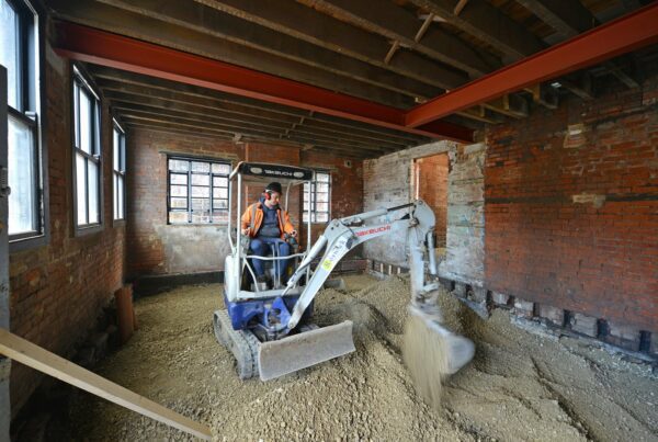 Construction worker in appropriate PPE using a digger to dig up rubble in an old brick room of a warehouse