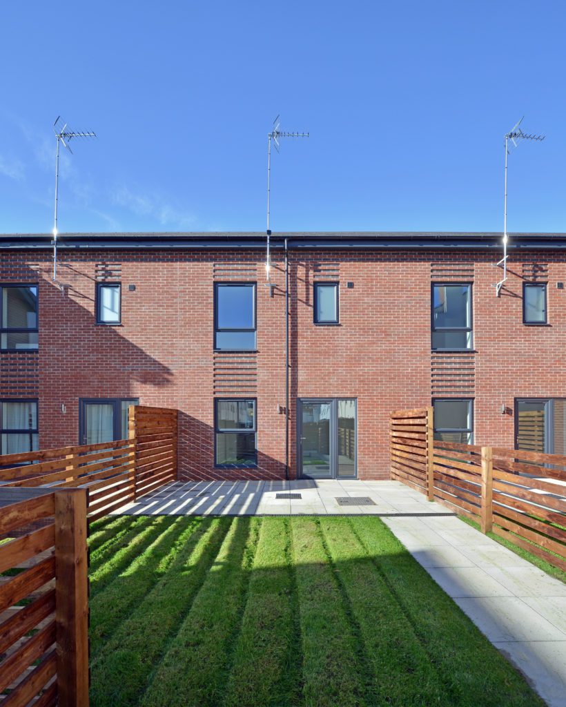 Rear facade of terraced, red brick residential buildings, with fenced back gardens.
