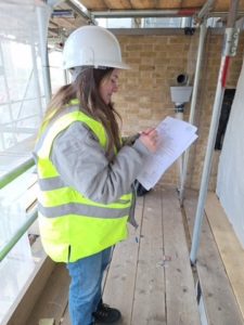 Georgia in a hard hat with a clip board taking notes on a construction site